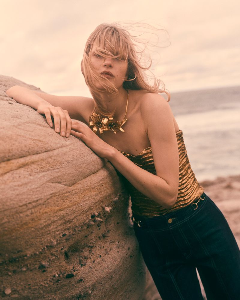 Adult female model with a blonde fringe poses next to a rock formation on a beach wearing a gold-colored strapless top and dark jeans.
