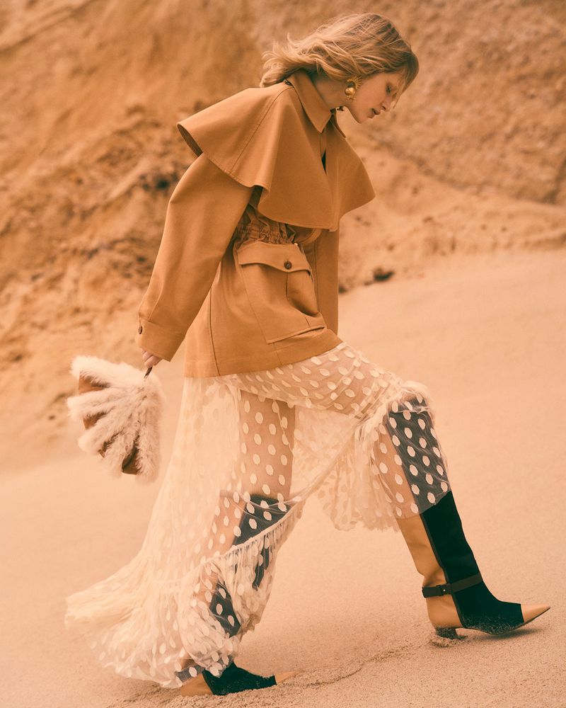 Fashionable woman poses in a stylish coat and polka dot dress, holding a feathered bag in a desert setting with light orange color grading.