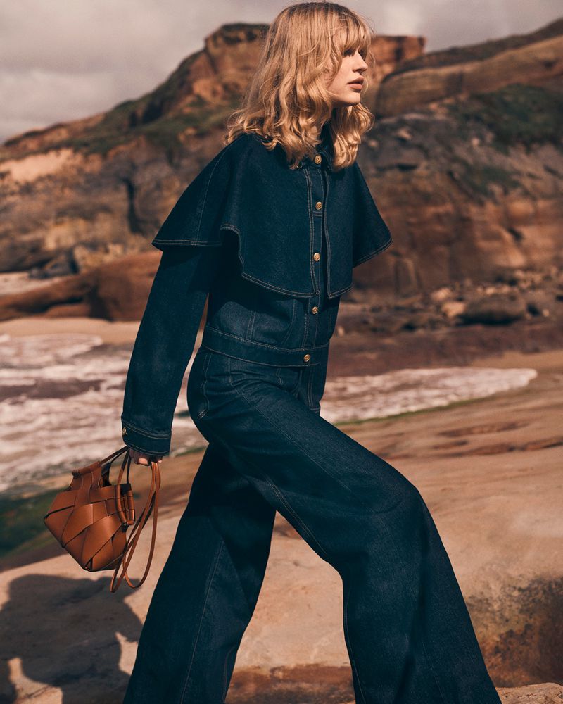 Fashion shot of a woman on a rocky beach wearing a dark denim jumpsuit and cape, carrying a woven leather handbag in neutral colors.