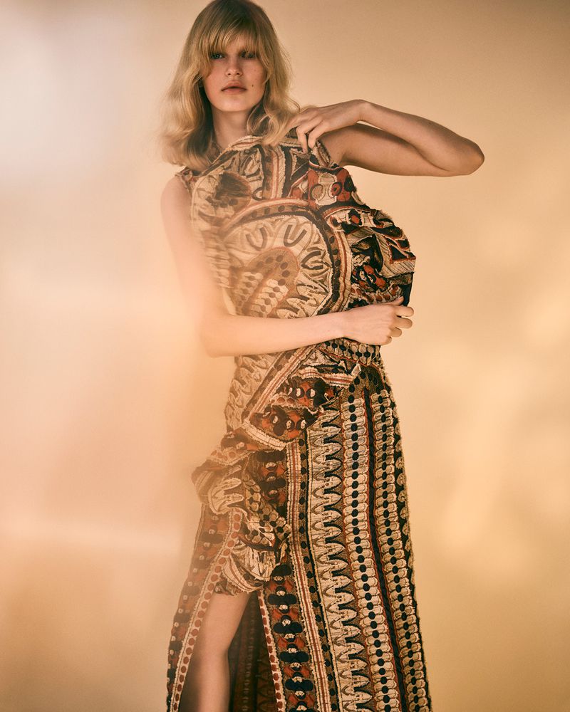 A blonde woman in a patterned brown and black sleeveless maxi dress holds the dress fabric in a studio with a blurred background.