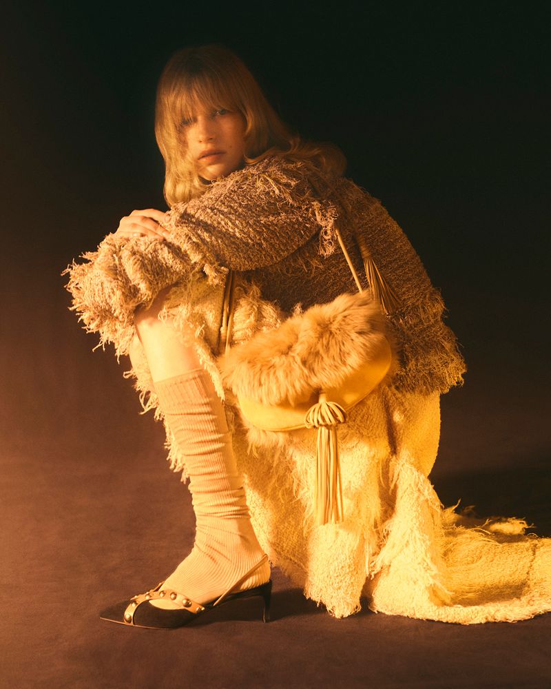 A blonde woman in a knitted outfit and yellow purse crouches in a studio setting with a dark backdrop, creating a fashionable and styled image.