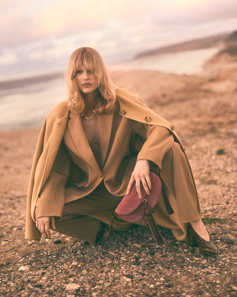 A blonde woman in a tan pantsuit crouches on a rocky beach with a burgundy suede bag, wearing dark flats; a body of water is in the background.