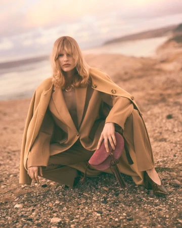 A blonde woman in a tan pantsuit crouches on a rocky beach with a burgundy suede bag, wearing dark flats; a body of water is in the background.