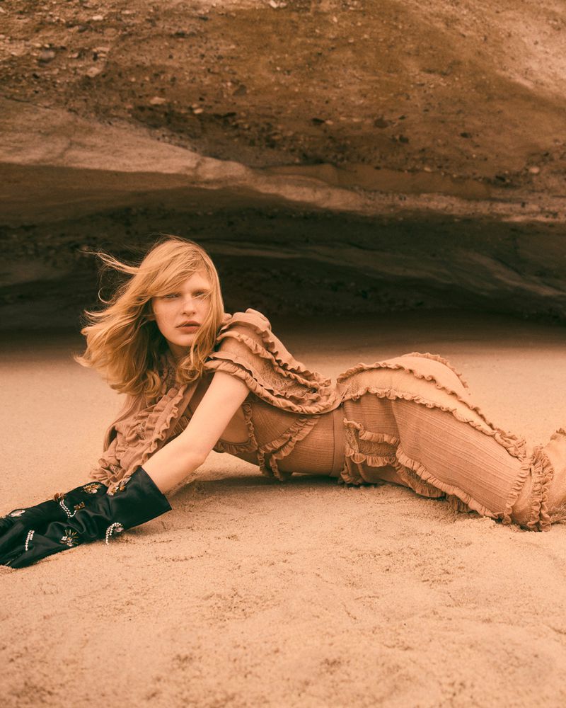 Woman in a beige dress and black gloves lies on a sandy beach with a rock formation in the background in a fashion editorial shoot.