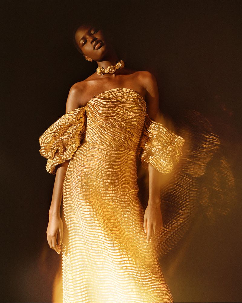 Striking full shot of an adult African female fashion model in a shimmering gold off-the-shoulder gown with statement gold choker against a dark background.