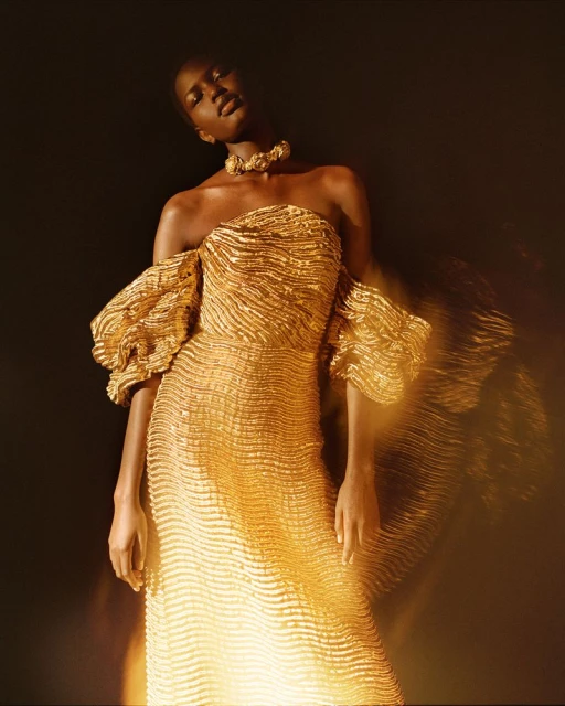 Striking full shot of an adult African female fashion model in a shimmering gold off-the-shoulder gown with statement gold choker against a dark background.