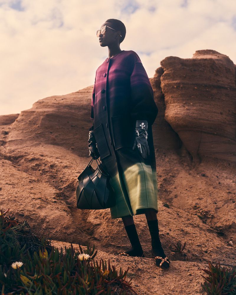 Stylish woman in sunglasses, coat, and gloves posing outdoors against a rocky landscape carrying a structured woven bag.