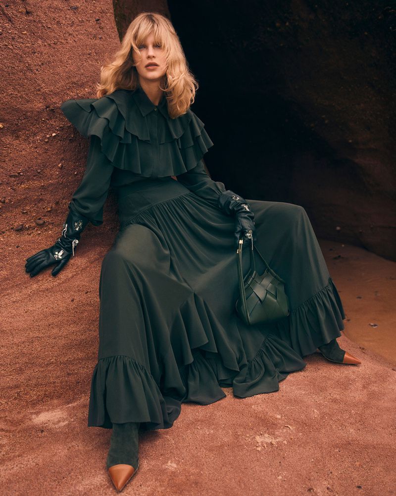 A stylish adult female poses in a long green ruffle dress, gloves, and boots on a red rock surface, with a small green purse in a natural cave.