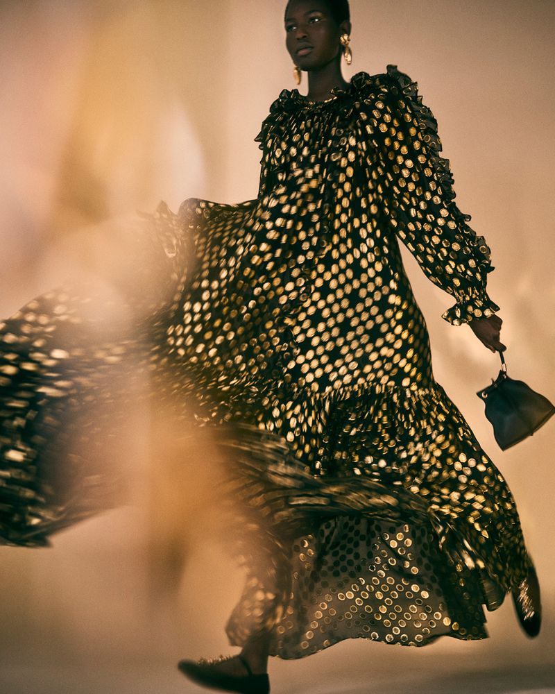 A Black female model in a flowing black and gold polka dot dress, holding a small black purse in a studio setting.