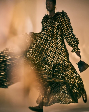 A Black female model in a flowing black and gold polka dot dress, holding a small black purse in a studio setting.