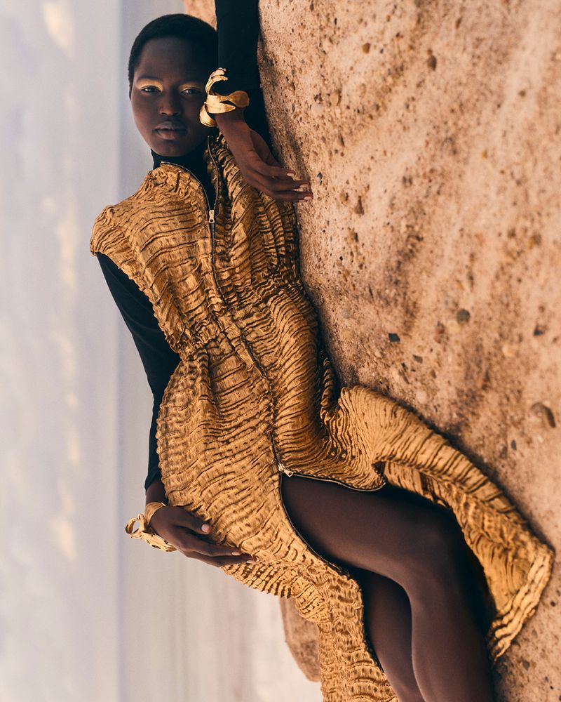 Woman in gold dress poses against rock formation with lake in background. Dark-skinned model in sleeveless dress with gold jewelry.