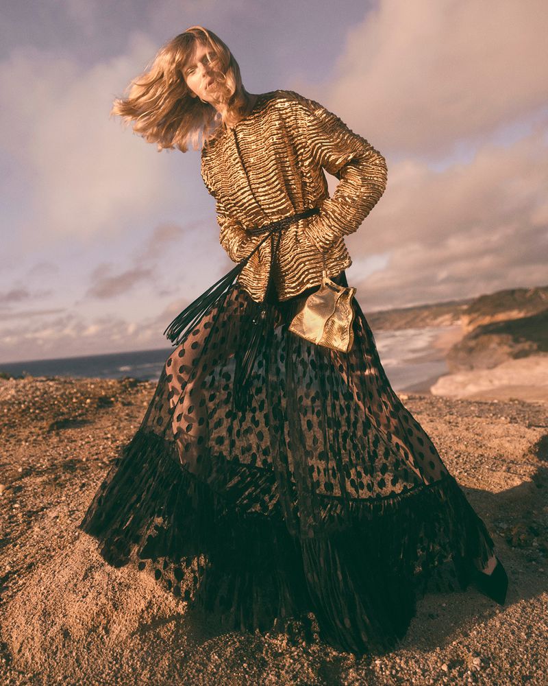 A woman in a shimmering gold jacket and a black spotted skirt stands on a sandy hilltop near a beach with waves and cloudy skies.
