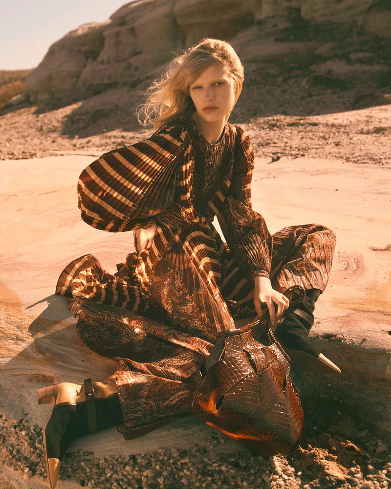 A blond model poses in a pleated dress and boots in an outdoor desert landscape with rocks and sand, holding a unique handbag.