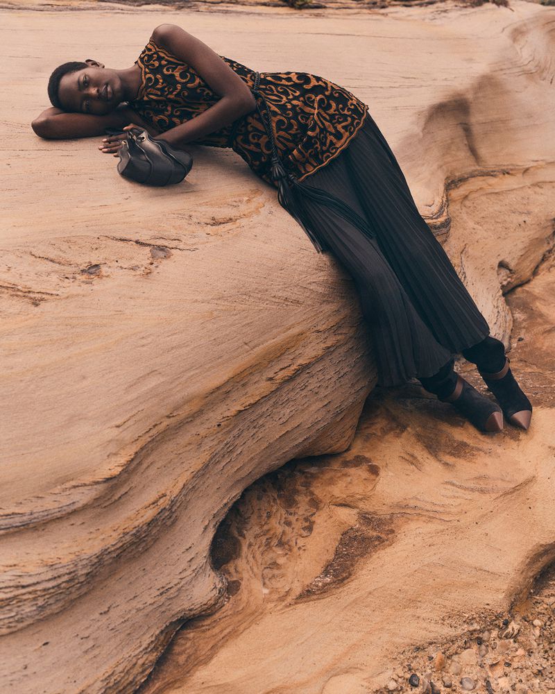 Fashion shot of an African woman in patterned sleeveless top and pleated skirt lying on tan rock formation, holding a small purse.