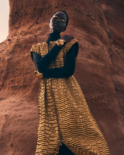 A dark-skinned model in a textured gold sleeveless dress poses against a red rock formation in a fashion editorial shoot.