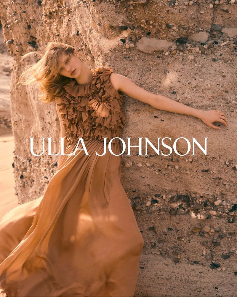 A woman in an earth-toned dress poses against a textured wall in natural light for a fashion editorial; Ulla Johnson logo visible.