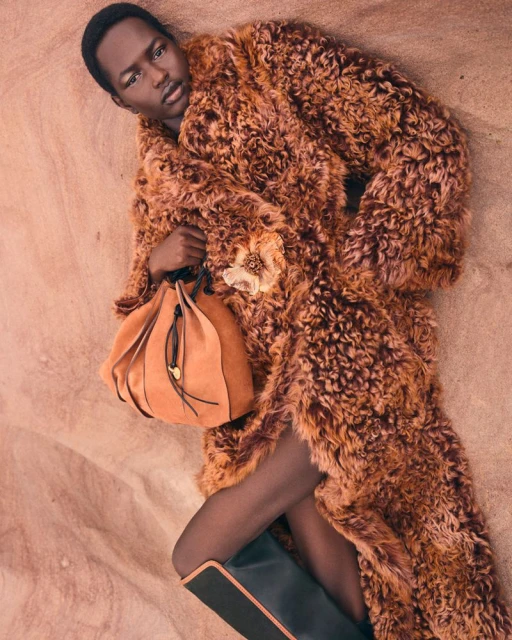 Editorial fashion shot of a Black model in a fuzzy brown coat, lying on a textured brown surface, holding a brown suede bucket bag.