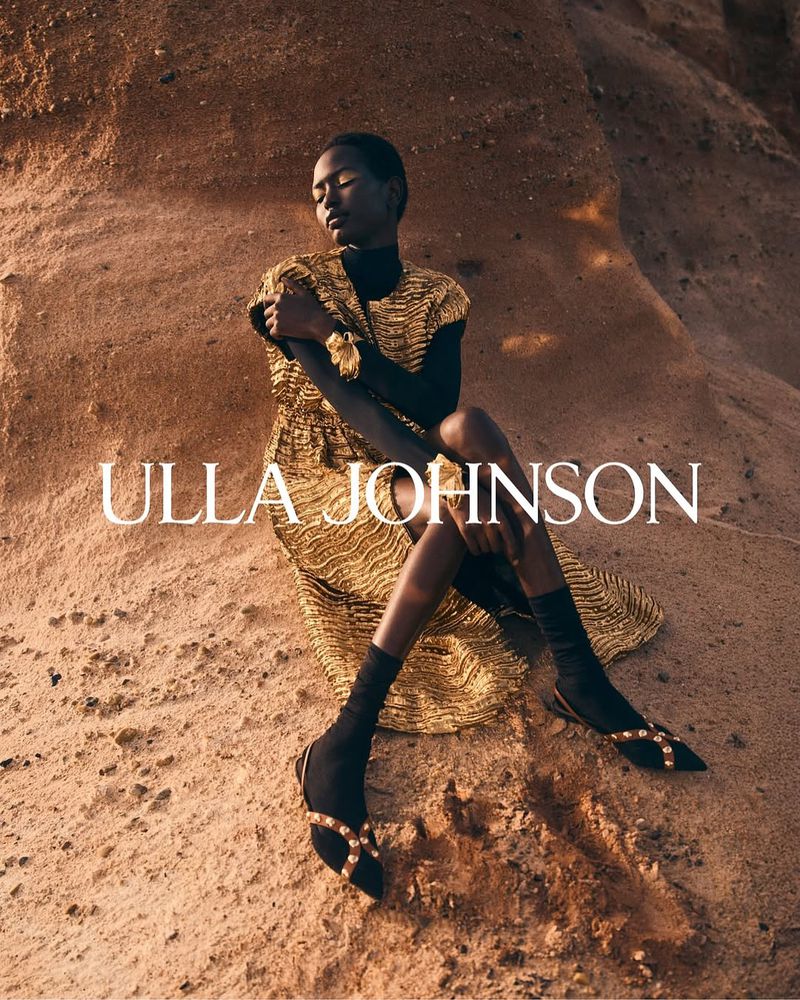 Woman in gold dress and black flats poses in a desert landscape. Fashion-forward attire in a natural setting with subdued, warm lighting.