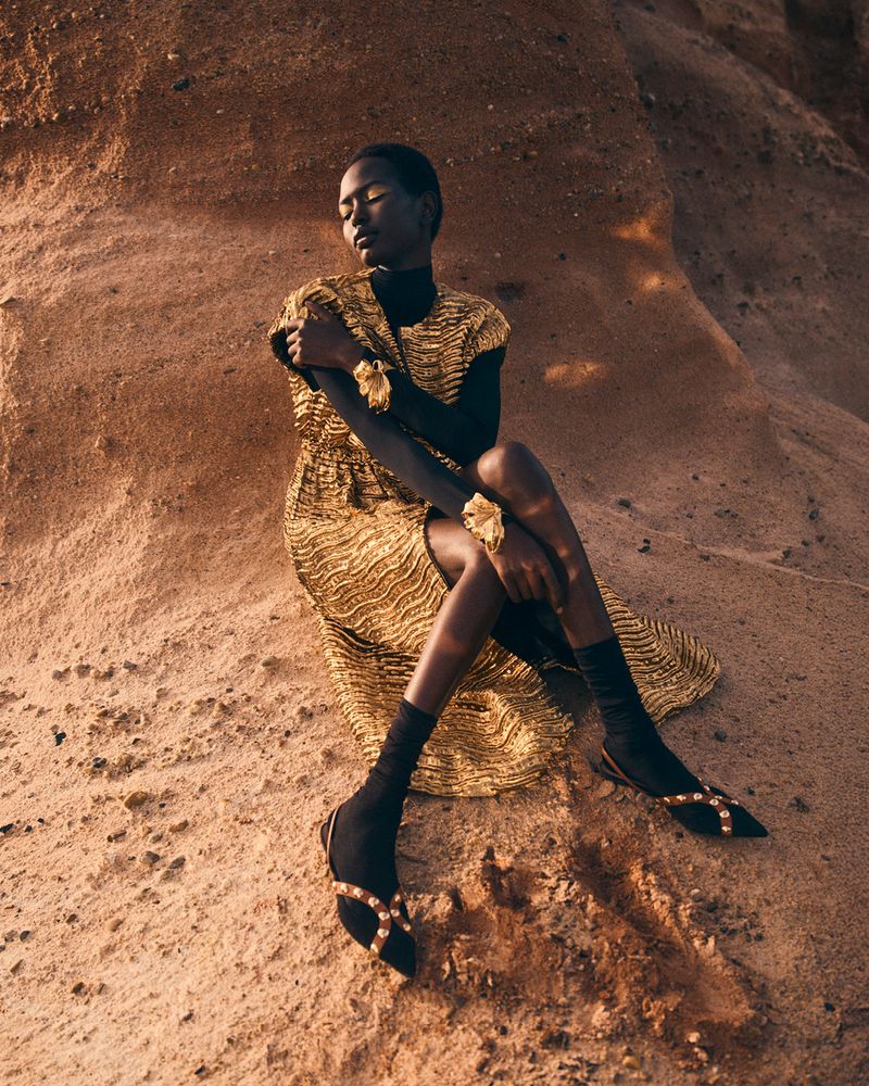 A young Black woman in a gold dress with black turtleneck, sitting in a desert landscape, fashionably posed, eyes closed, against rock formations.
