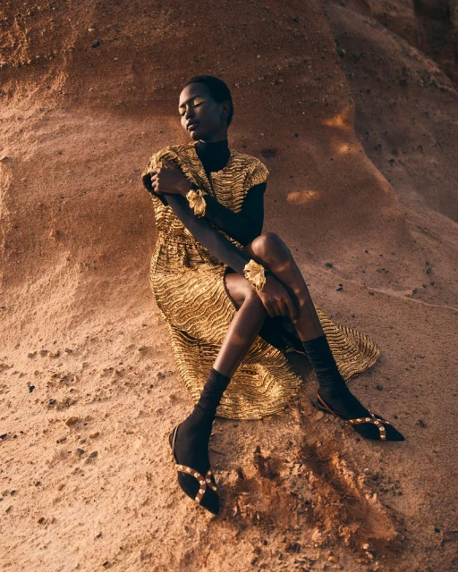 A young Black woman in a gold dress with black turtleneck, sitting in a desert landscape, fashionably posed, eyes closed, against rock formations.