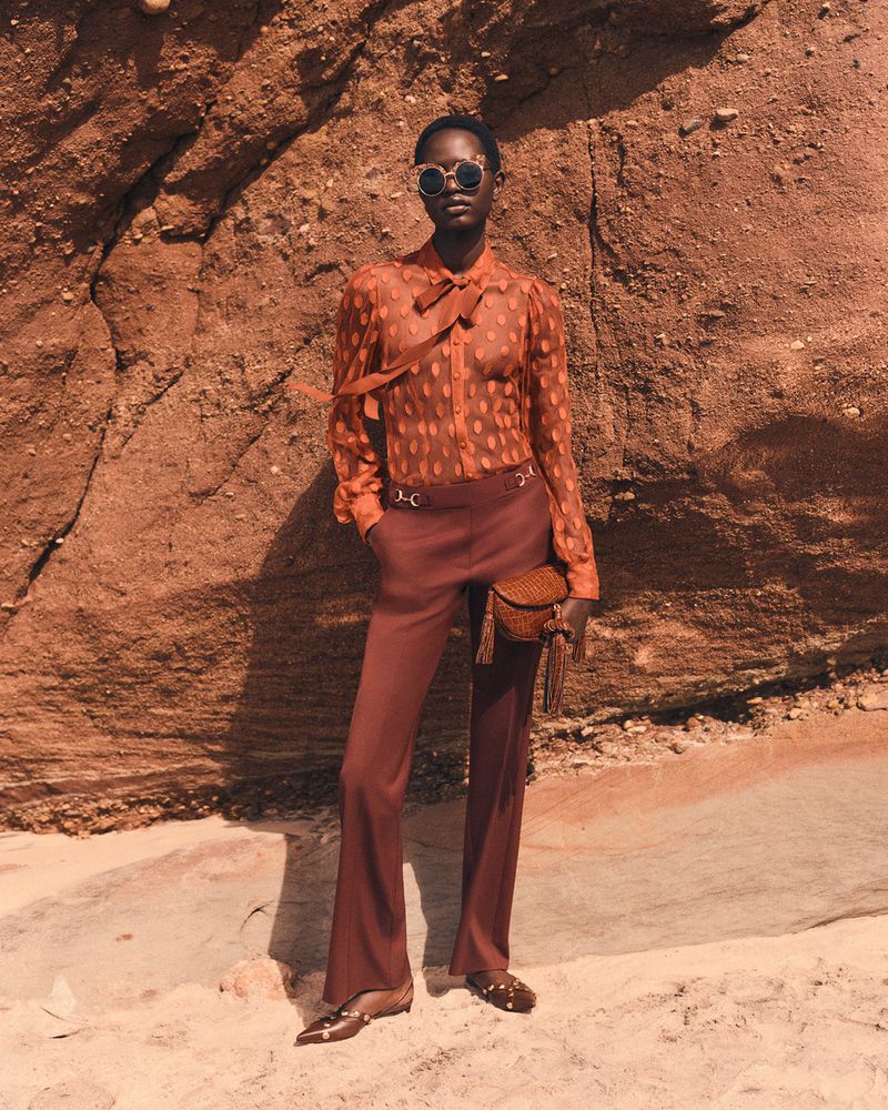 A woman in retro-style clothing poses against a textured, red-toned rock face in a fashion editorial shot with warm, diffused lighting.