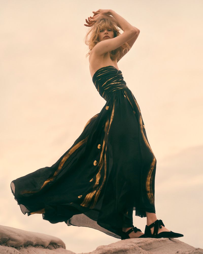 A woman in a flowing black and gold dress stands on rocks against a soft sky, photographed in an outdoor fashion editorial style.