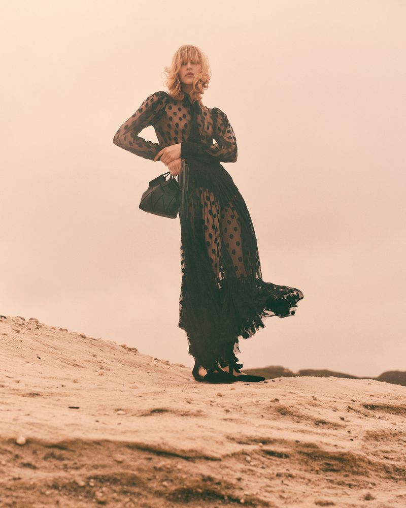A woman in a black polka dot dress poses on a sandy dune, holding a small black purse, with a pale orange sky in the background.