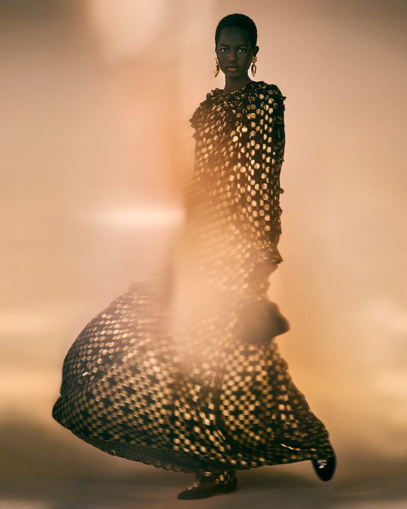 Fashion editorial shot featuring a Black female model wearing a black and gold dress, posing in a diffused, warm-toned background.