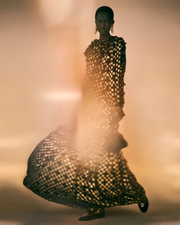 Fashion editorial shot featuring a Black female model wearing a black and gold dress, posing in a diffused, warm-toned background.