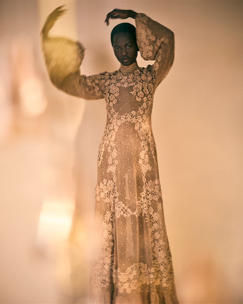 A graceful Black model poses in an elegant, long, beige lace dress with puffed sleeves, her hands raised, set against a warm, soft background.