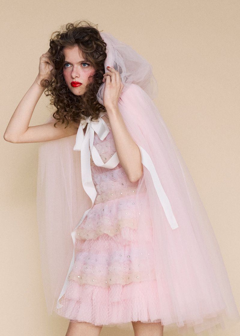 Young woman posing in pink tulle dress and hooded cape with white ribbon, soft studio lighting, light peach background, fashion photography.