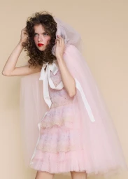 Lily Nova wearing a tiered pink tulle dress and matching hooded cape tied with a white ribbon bow against a beige backdrop.