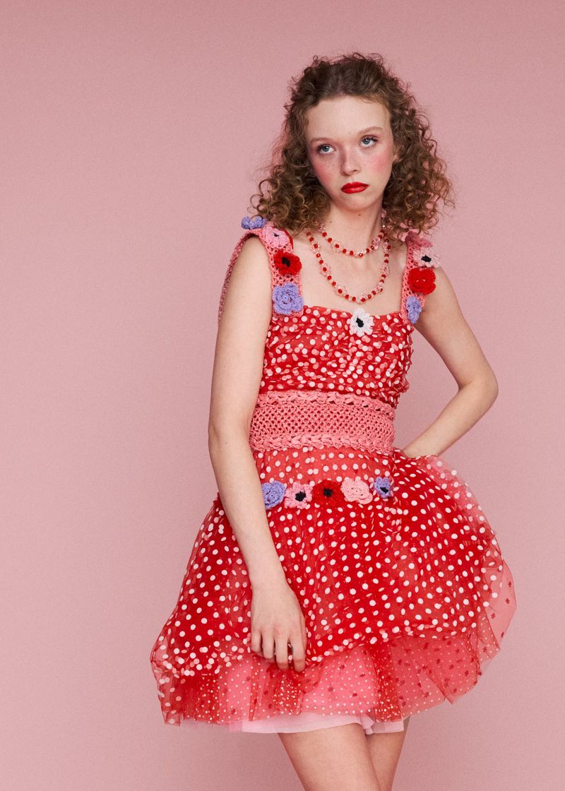 Model Lily Nova wears a red polka-dot tulle mini dress adorned with crochet flowers against a solid pink backdrop for Ryan LO.