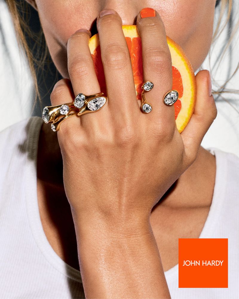 Close-up of a person holding a grapefruit slice, showcasing two gold rings with clear stones, studio lighting, and an orange logo.