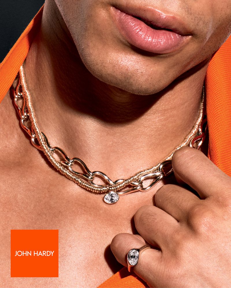 Close-up of a man wearing a gold chain necklace with a gemstone pendant, and a ring, in a studio shot with a dark background.