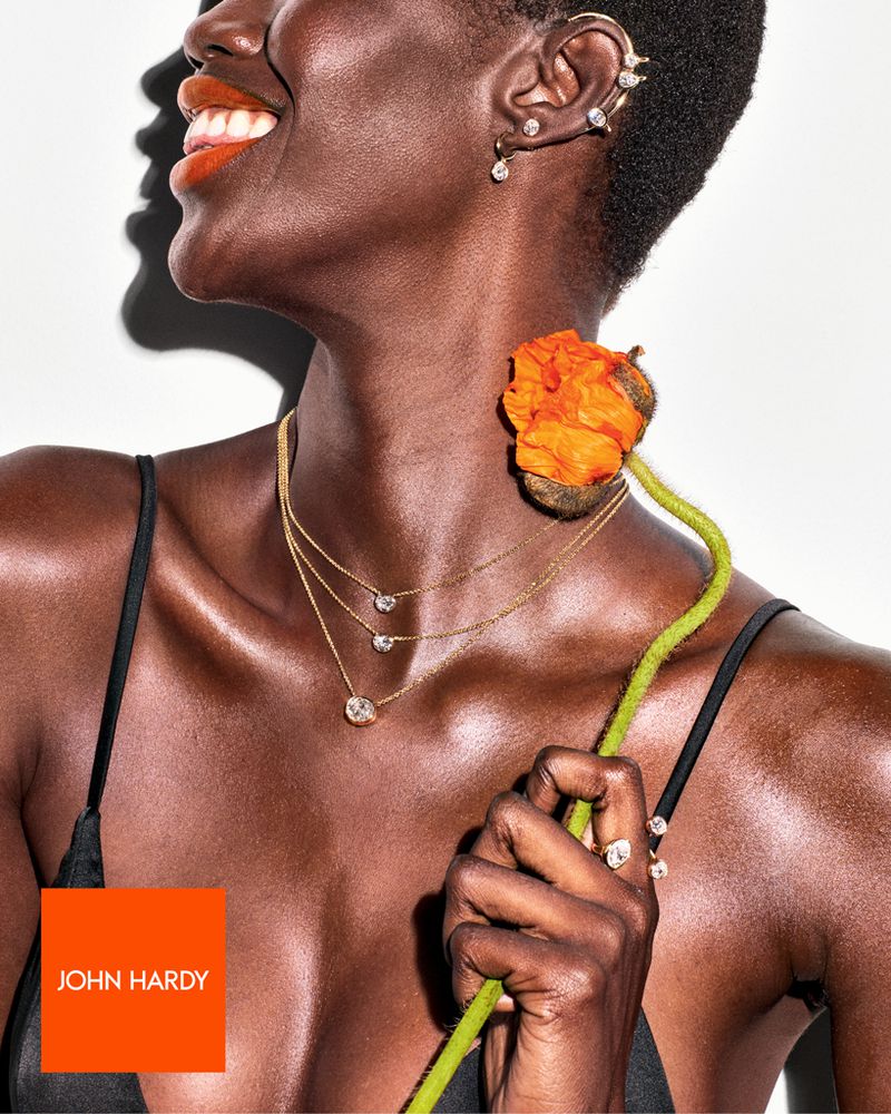 Dark-skinned woman poses in a close-up fashion shot wearing gold jewelry, including a triple necklace and ear piercings, with a red-orange flower.