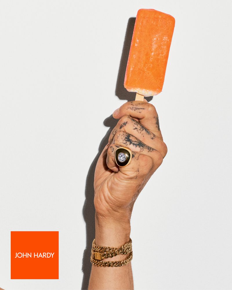 A hand holds an orange popsicle, adorned with gold John Hardy bracelets and ring against a plain white background in a studio setting.