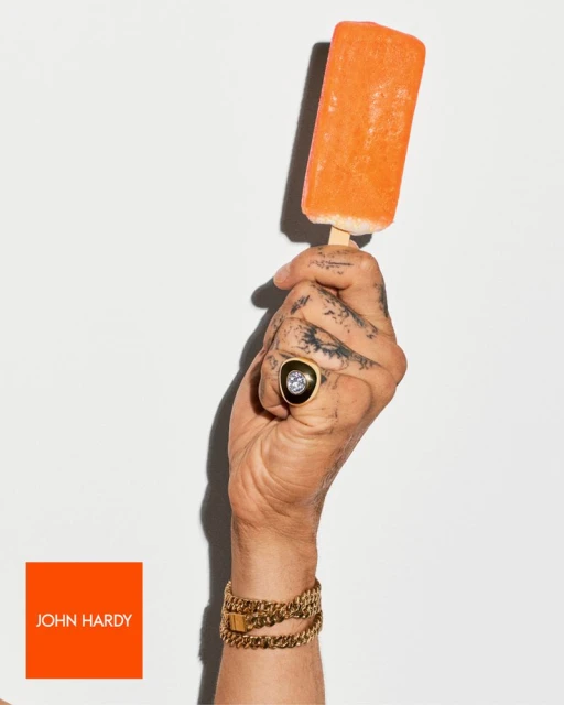 A hand holds an orange popsicle, adorned with gold John Hardy bracelets and ring against a plain white background in a studio setting.