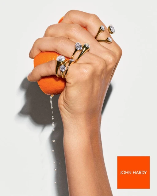 Hand squeezing an orange, with water droplets, fingers adorned with gold gemstone rings, John Hardy logo in the bottom right corner.