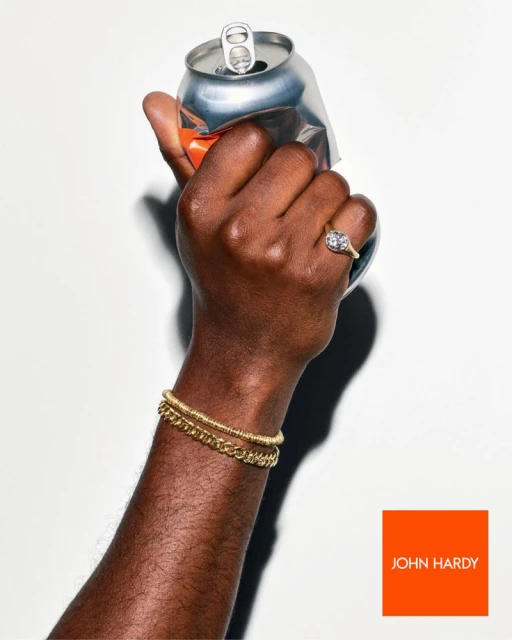 Elevated studio shot of a dark-skinned hand crushing a soda can, adorned with gold bracelets and a diamond ring, against a white background.