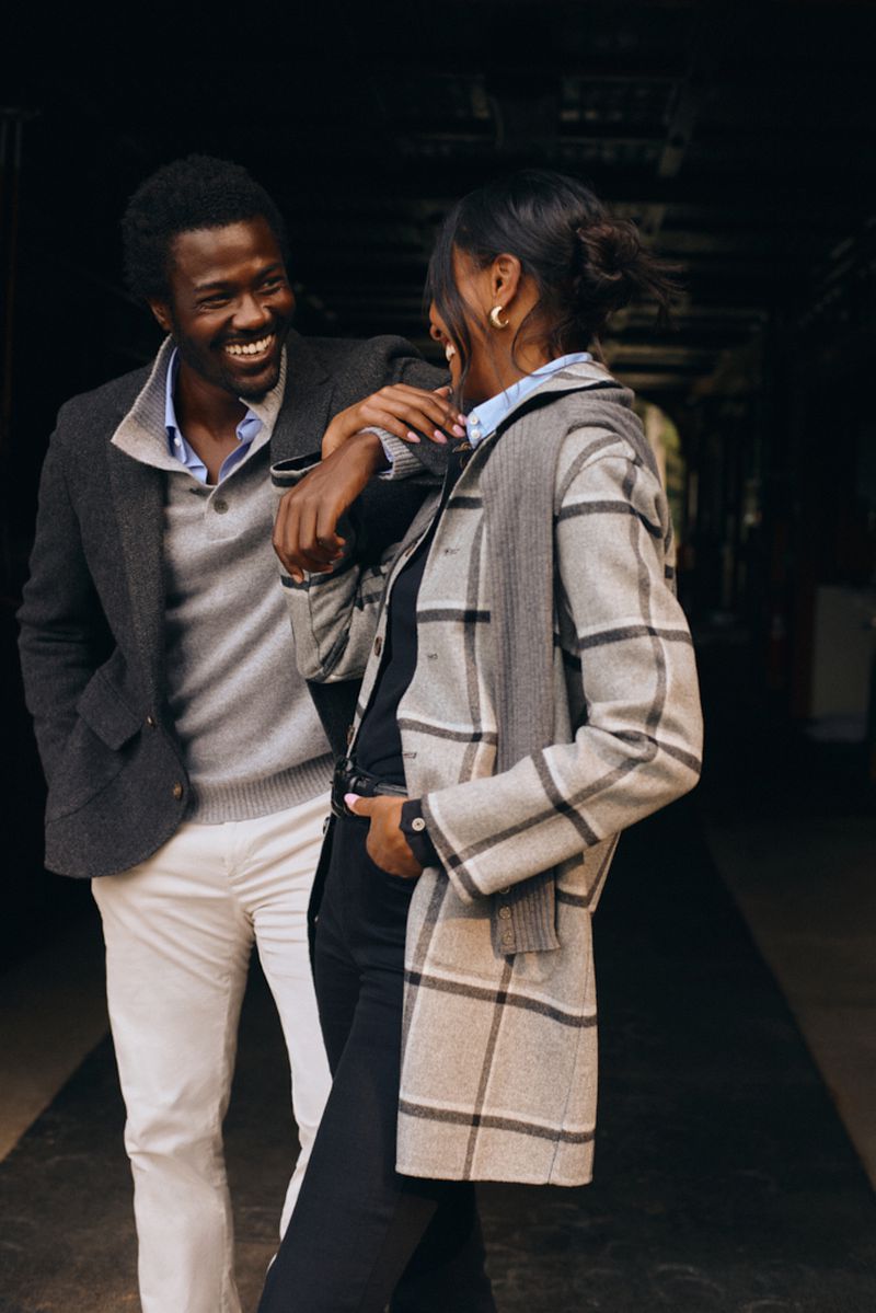 A joyful African-American couple is posing outdoors. The man is wearing a sport coat with white pants. The woman wears a plaid coat.