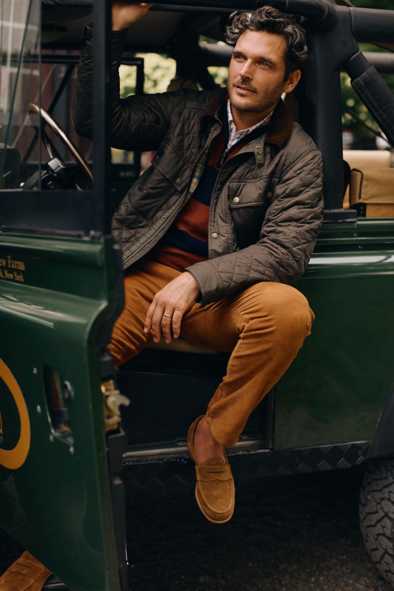 Handsome adult man wearing a quilted jacket and brown pants sitting in an olive green off-road vehicle in a natural outdoor environment.