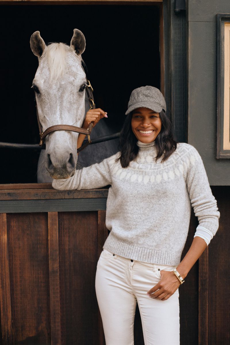 A woman smiles next to a grey horse in a stable, wearing a patterned sweater, white pants, and a cap. The horse is wearing a bridle.