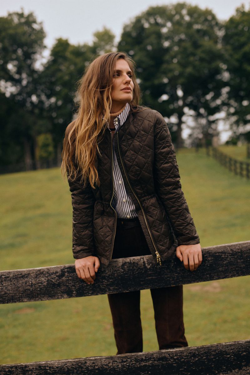 Woman in quilted jacket poses outdoors by wooden fence. She wears a striped shirt and dark trousers in a natural setting.