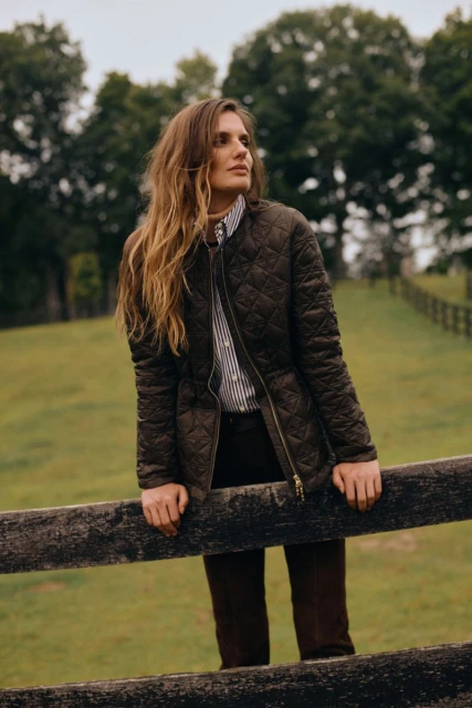 Woman in quilted jacket poses outdoors by wooden fence. She wears a striped shirt and dark trousers in a natural setting.
