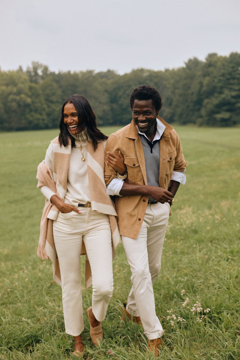 A smiling couple walks in a grassy field wearing cream and tan fall fashions, with trees in the background under an overcast sky.