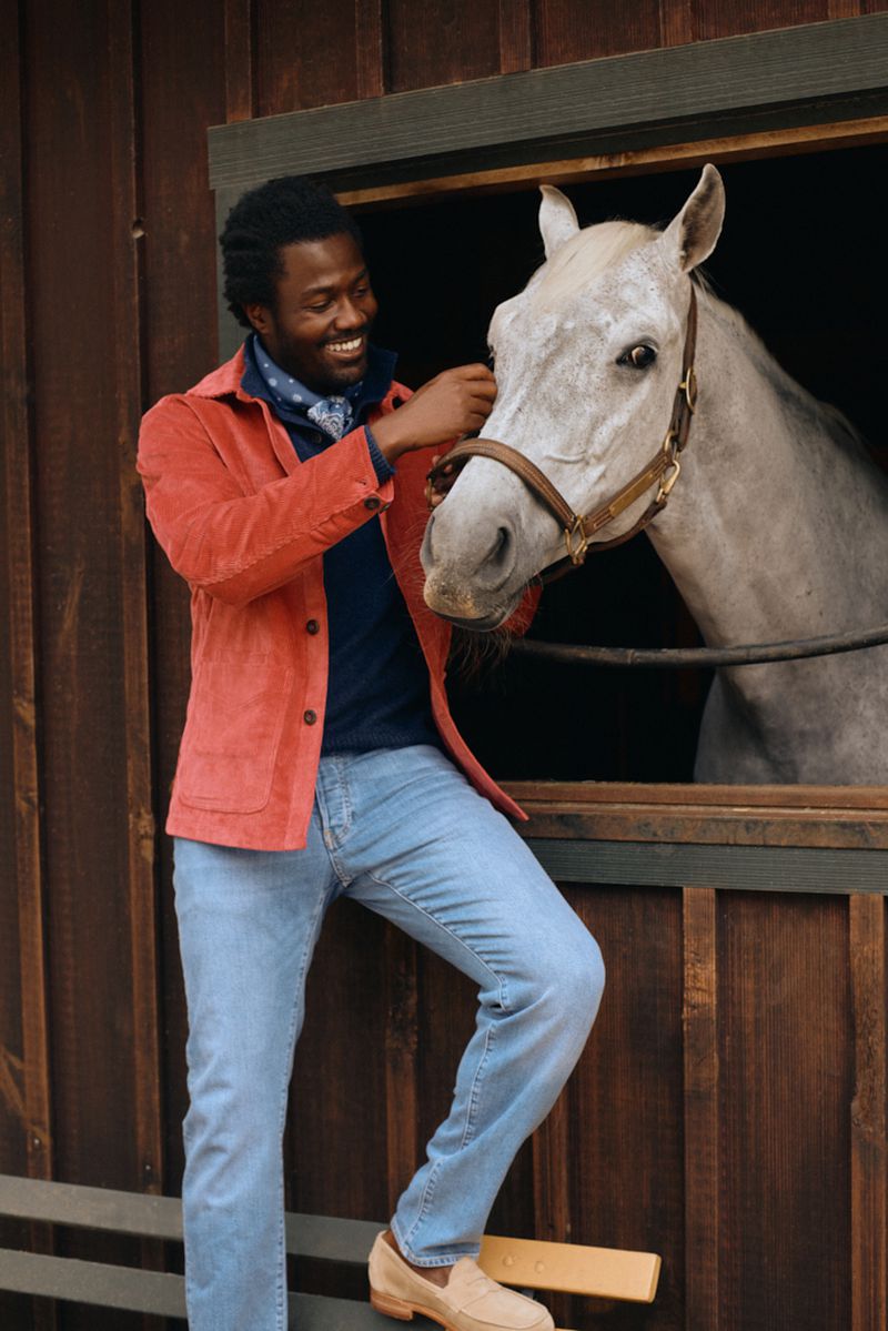 An African-American man stands beside a white horse in a stable, smiling and looking at the horse, wearing jeans and a red corduroy jacket.
