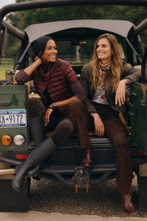 Two women in autumn fashion sit on the back of a green SUV with an open top, posing in a rural setting with trees.