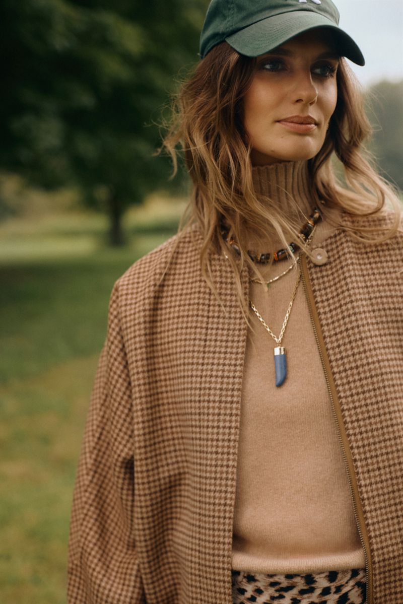 Brunette woman in green baseball cap and brown houndstooth coat stands outdoors wearing layered gold necklaces and neutral turtleneck.