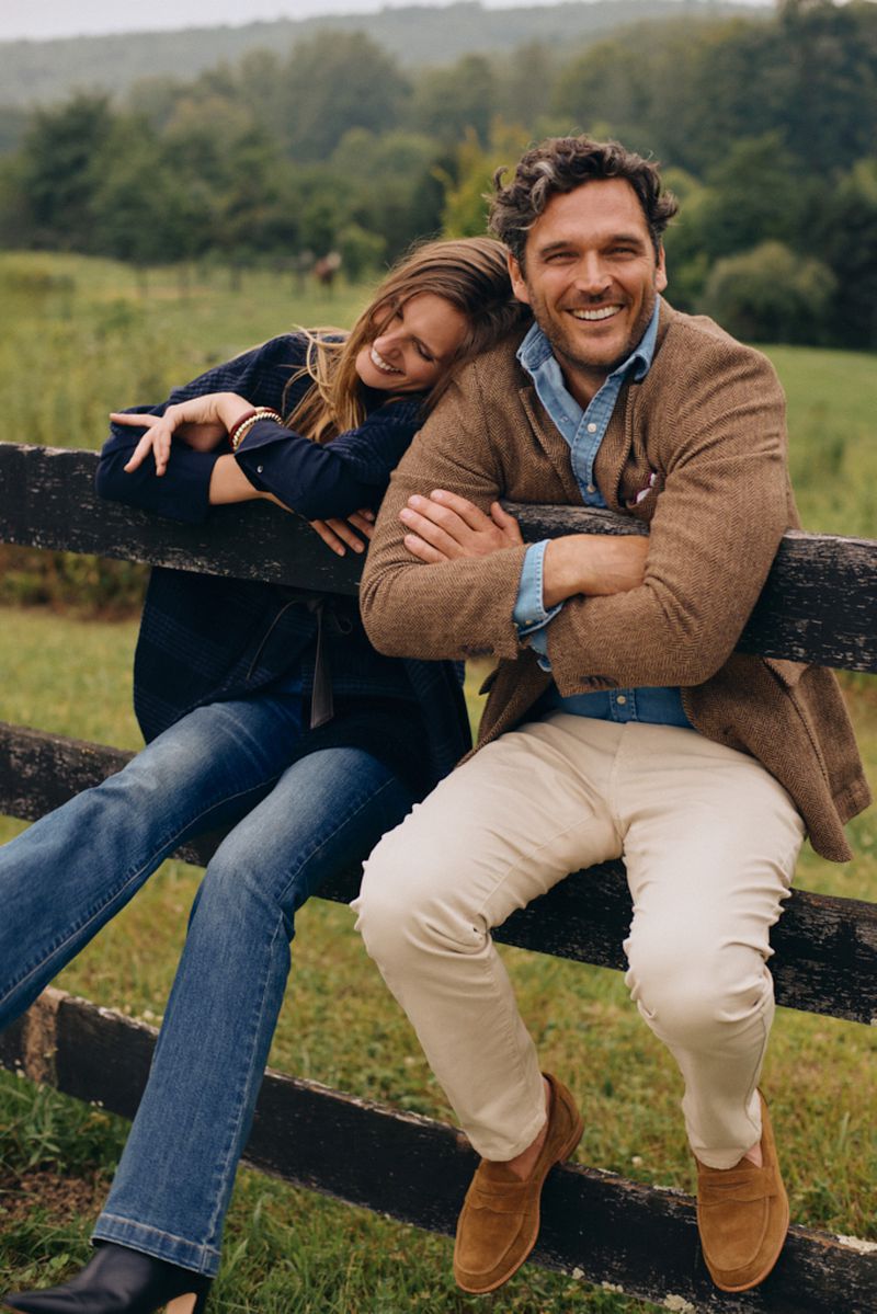 A smiling couple sits on a wooden fence in a rural setting, enjoying the outdoors. The man and woman wear smart casual clothing.